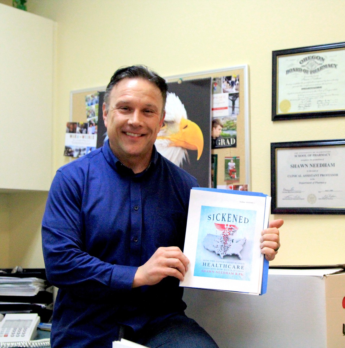Moses Lake pharmacist Shawn Needham, sitting just outside his office, holds up a printout of the cover of his book “Sickened: How the Government Ruined Healthcare and How to Fix it.”