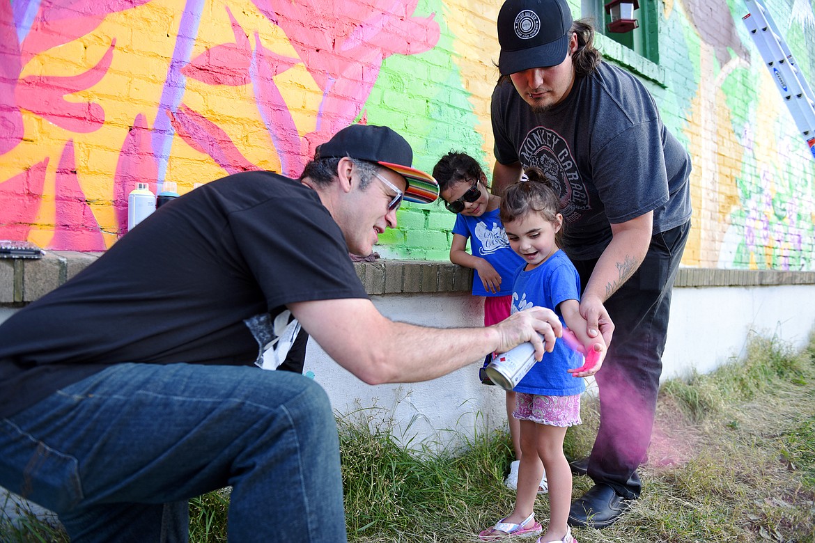 Artist Thomas Valencia spray-paints the hand of Raven Stanley as her sister, Violet, and father, Jordan, watch below a mural Valenica is creating along West Center Street and Fifth Avenue West in Kalispell on Tuesday, Aug. 4. Valencia is asking for the community's help in filling the white horizontal panel with hand prints, as long as you don't mind getting spray paint on your hand. (Casey Kreider/Daily Inter Lake)