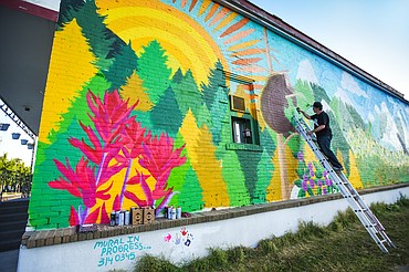 Thomas Valencia, of Creston, works on a mural along West Center Street at Fifth Avenue West in Kalispell on Tuesday, Aug. 4. Valencia began working on the project about 10 days ago and used GoFundMe to crowdsource money for primer paint, low-pressure spray paint cans for the detail work and additional supplies. Valencia also seeks the community's help with filling the lower white panel of the mural with painted hand prints, as long as you don't mind getting a little spray paint on your hands. He primarily works on the project during the evening hours, which he received permission to complete from the building owner, Valley Linen, and the city of Kalispell. The mural features snowcapped mountains, rushing rivers and streams, wildlife, forests and flowers and will transition from a daylight to a nighttime scene from left to right. The mural will span an entire block when complete. (Casey Kreider/Daily Inter Lake)