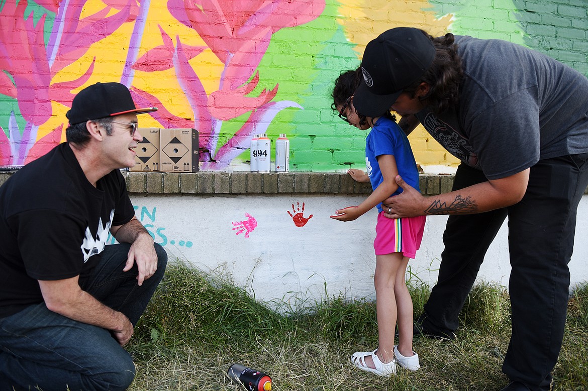 Artist Thomas Valencia, left, watches as Jordan Stanley, right, helps his daugther, Violet, press her painted hand print onto a wall below a mural Valencia is creating along West Center Street and Fifth Avenue West in Kalispell on Tuesday, Aug. 4. Valencia is asking for the community's help in filling the white horizontal panel with hand prints, as long as you don't mind getting spray paint on your hand. (Casey Kreider/Daily Inter Lake)