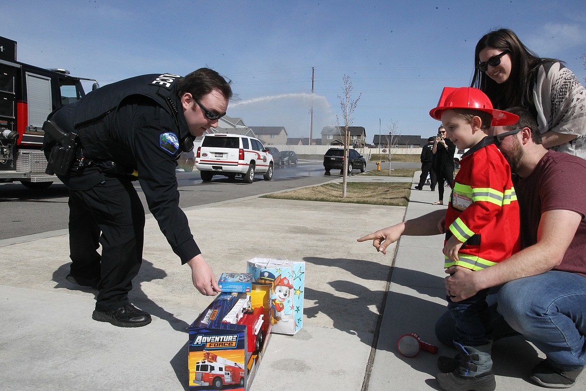 BILL BULEY/Press Rathdrum Police officer Scott Kennedy presents a gift to Lukas Petroskie, standing with dad Matt and mom Hannah, as they watch the Northern Lakes Fire District units and police vehicles pass their Rathdrum home Wednesday afternoon to celebrate the boy's fourth birthday.