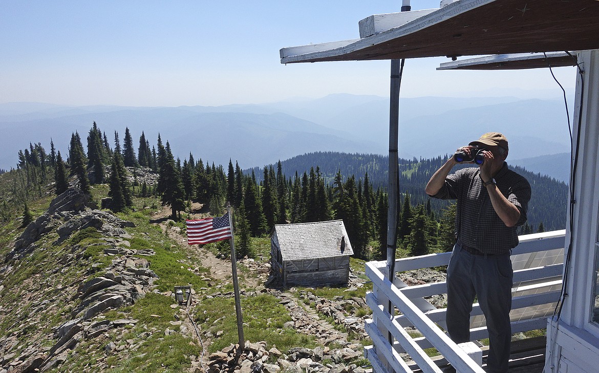 In this Wednesday, July 18, 2018, photo, Tom VandeWater stands at the railing of the Coolwater Fire Lookout and looks through his binoculars in the Nez Perce-Clearwater National Forests near Lowell, Idaho. VandeWater, from Canton, N.Y., has staffed the lookout each summer for many years for the U.S. Forest Service. At left is the previous lookout structure, which dates to the 1930s. (AP Photo/Ted S. Warren)