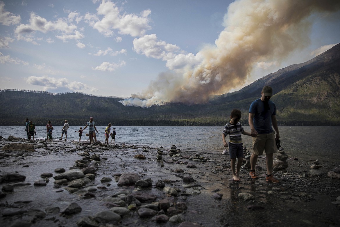 FILE - In this Sunday, Aug. 12, 2018 file photo provided by the National Park Service, people walk along the shore near Lake McDonald Lodge as the Howe Ridge Fire burns in Glacier National Park, Mont. Wildfires that have kept portions of Glacier National Park closed for two weeks are scrambling visitors&#146; plans and prompting some to cancel their trips. Much of Glacier&#146;s famous Going-to-the-Sun-Road has been closed since August 12 due to a fire that&#146;s burned more than 19 square miles (50 square kilometers). (National Park Service via AP, File)