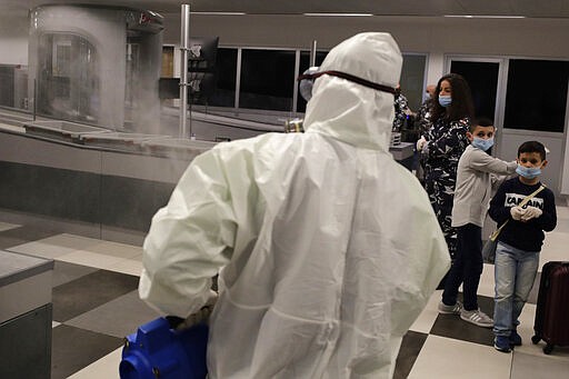 Passengers wear protective masks while waiting for their plane look to a worker wearing protective gear sprays disinfectant as a precaution against the coronavirus outbreak, in the departure terminal at the Rafik Hariri International Airport, in Beirut, Lebanon, Thursday, March 5, 2020. The novel coronavirus has infected more than 80,000 people globally, causing around 2,700 deaths, mainly in China. (AP Photo/Hassan Ammar)