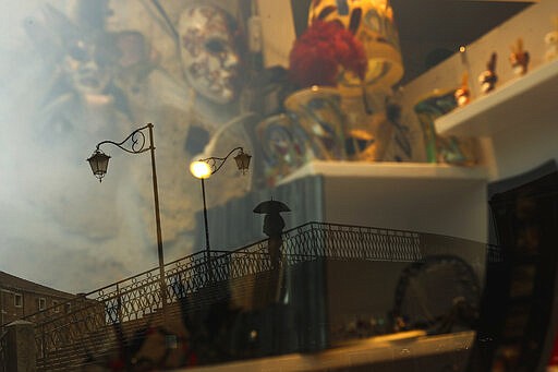 A man, reflected in a carnival masks shop display window, crosses a bridge at the Murano island in Venice, Italy, Monday, March 2, 2020. The Carnival period in Venice usually marks the start of peak season in one of the world's most visited cities, with hordes of tourists piling onto vaporettos to cruise the Grand Canal, strolling through cobble-stoned streets and lingering in picturesque cafes. Venice in the time of coronavirus, though, is a shell of itself, with empty piazzas, shuttered basilicas and gondoliers idling their days away. (AP Photo/Francisco Seco)