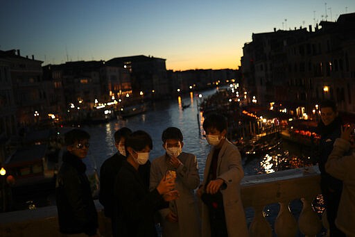 Tourists wearing protective masks pose for a photograph at the Rialto bridge as the sun sets in Venice, Italy, Friday, Feb. 28, 2020. Venice in the time of coronavirus is a shell of itself, with empty piazzas, shuttered basilicas and gondoliers idling their days away. The city has remained quiet ever since the outbreak, with only intrepid tourists wearing a different type of mask _ surgical _ remaining and taking advantage of a city that otherwise would be jammed. (AP Photo/Francisco Seco)