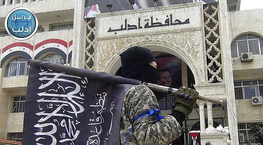 FILE - In this file photo posted on the Twitter page of Syria's al-Qaida-linked Nusra Front on March 28, 2015, a fighter from Syria's al-Qaida-linked Nusra Front holds his group flag as he stands in front of the governor building in Idlib province, north Syria. Intermingled among 3 million civilians under siege in the Syrian government's assault on the last opposition stronghold are tens of thousands of al-Qaida-linked fighters and other militants who came from around the world to take part in the country's civil war. (Al-Nusra Front Twitter page via AP, File)