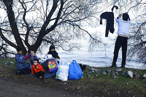 A migrant who arrived yesterday to Greece from Turkey dries his clothes as a family sit under a tree at the village of Skala Sikaminias, on the Greek island of Lesbos on Friday, March 6, 2020. Thousands of refugees and other asylum-seekers have tried to enter Greece from the land and sea in the week since Turkey declared its previously guarded gateways to Europe open. (AP Photo/Alexandros Michailidis)