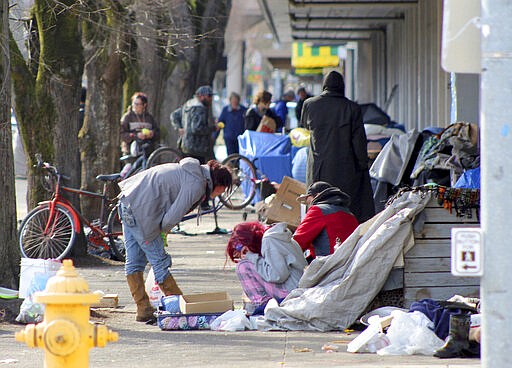 In this Tuesday, March 3, 2020 photo, homeless people crowd a sidewalk in downtown Salem, Ore., where they have set up a makeshift camp. Experts say that the homeless, who often have health and substance-abuse problems, are exposed to the elements and do not have easy access to hygiene, are more vulnerable to the coronavirus. Some cities are making provisions so the homeless who contract the virus have a place to recover without spreading the infection further. (AP Photo/Andrew Selsky)