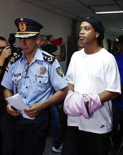 Former soccer star Ronaldinho walks handcuffed and escorted by a police officer to declare to judge Clara Ruiz Diaz at Justice Palace in Asuncion, Paraguay, Saturday, March 7, 2020. Ronaldinho and his brother Roberto were alleged to enter to the country with a false Paraguayan passport.(AP Photo/Jorge Saenz)