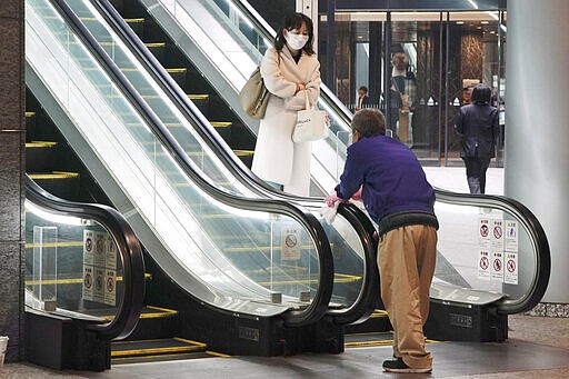 A woman wearing a mask as a precaution against a new coronavirus passes by a worker cleaning escalator handrails at a business building in Tokyo Monday, March 9, 2020. (AP Photo/Eugene Hoshiko)