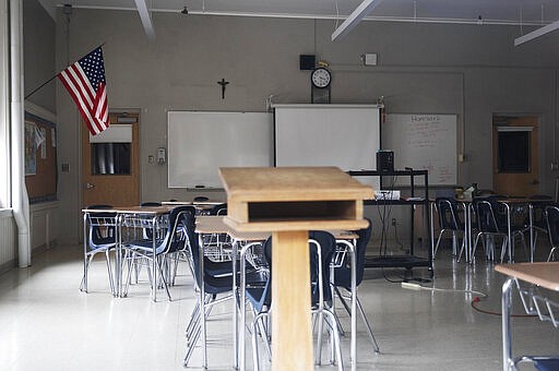 In this March 6, 2020, photo, a classroom is seen vacant through a window at Saint Raphael Academy in Pawtucket, R.I., as the school remains closed following a confirmed case of the coronavirus. As a growing number of schools around the country close their doors because of the new coronavirus, they are confronted with the dilemma of whether to move classes online and run the risk of leaving behind the many students who don't have internet or computers at home, or parents with flexible work schedule. (AP Photo/David Goldman)