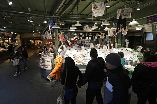 In this March 6, 2020 photo, a handful of people watch as Colin Kiplinger, center-left, tosses a salmon to a colleague behind the counter as he works at Pike Place Fish Market in Seattle. Foot traffic and crowds at the Pike Place Market, which is popular with tourists and locals alike, have slowed as some workers stay home and work remotely, and some tourists cancel trips over worry about the COVID-19 coronavirus. (AP Photo/Ted S. Warren)