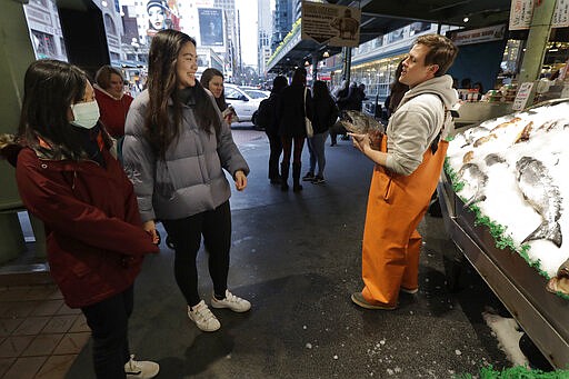 In this March 6, 2020 photo, a woman wears a mask as she joins a few others in watching Colin Kiplinger, right, as he works at Pike Place Fish Market in Seattle. Foot traffic and crowds at the Pike Place Market, which is popular with tourists and locals alike, have slowed as some workers stay home and work remotely, and some tourists cancel trips over worry about the COVID-19 coronavirus. (AP Photo/Ted S. Warren)