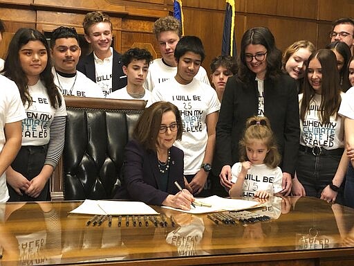 Oregon Gov. Kate Brown signs an executive order on Tuesday, March 10, 2020, ordering the state to lower greenhouse gas emissions, as students look on in Salem, Ore. In an end run around Republican legislators, Oregon's Democratic governor ordered the state on Tuesday to lower greenhouse gas emissions, directing a state agency to set and enforce caps on pollution from industry and transportation fuels. (AP Photo/Andrew Selsky)