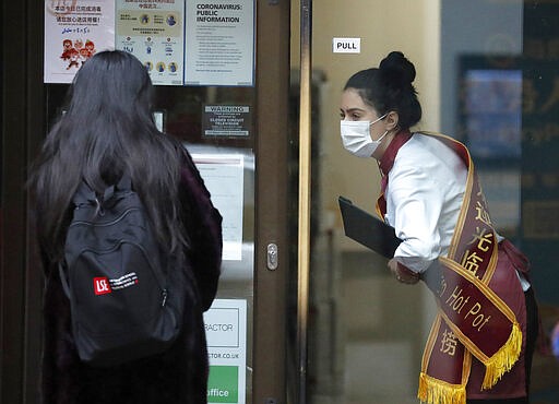 In this March 5, 2020, photo, a waiter of a Chinese restaurant wearing a face mask welcomes a customer in London. While white collar workers trying to avoid contagion can work from home or call in sick if they experience symptoms of the new virus, such precautions are not an options for the millions of waiters, delivery workers, cashiers, ride-hailing drivers, museum attendants and countless others who routinely come into contact with the public. (AP Photo/Frank Augstein)