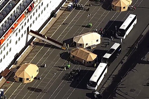 This photo taken from pool video provided by KGO-TV, shows passengers waiting to board buses after leaving the Grand Princess cruise ship at the Port of Oakland, Tuesday, March 10, 2020, in Oakland, Calif.  Passengers wearing masks trickled out of the Grand Princess and walked to the bottom of a ramp, where masked officials in yellow protective gear and blue plastic gloves took their temperature and led them to a tent for more screening before they lined up to board a bus to quarantine. (KGO-TV via AP, Pool)