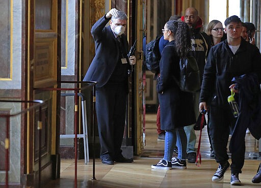 FILE - In this Thursday, March 5, 2020 file photo, an employee of the Palace of Versailles wearing a mask and gloves guides a tourist at the castel in Versailles, west of Paris. While white collar workers trying to avoid contagion can work from home or call in sick if they experience symptoms of the new virus, such precautions are not an options for the millions of waiters, delivery workers, cashiers, ride-hailing drivers, museum attendants and countless others who routinely come into contact with the public. (AP Photo/Michel Euler, File)