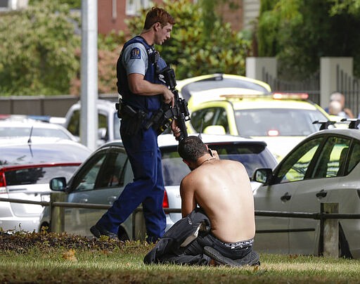 FILE - In this March 15, 2019, file photo, a man rests on the ground while speaking on his mobile phone as an armed police officer walks past near the Al Noor mosque following a mass shooting in central Christchurch, New Zealand. On Sunday, March 15, 2020, New Zealand will commemorate the 51 people who were killed in the attacks. (AP Photo/Mark Baker, File)