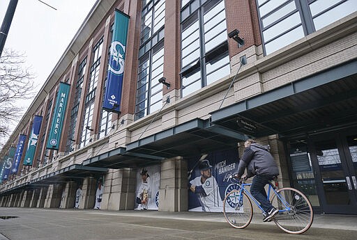 A bicyclist rides past T-Mobile Park, Wednesday, March 11, 2020, in Seattle, where baseball's Seattle Mariners plays home games. In efforts to slow the spread of the COVID-19 coronavirus, Washington State Gov. Jay Inslee announced a ban on large public gatherings in three counties in the metro Seattle area. That decision impacts the Seattle Mariners, Seattle Sounders, and the XFL's Seattle Dragons home games. (AP Photo/Stephen Brashear)