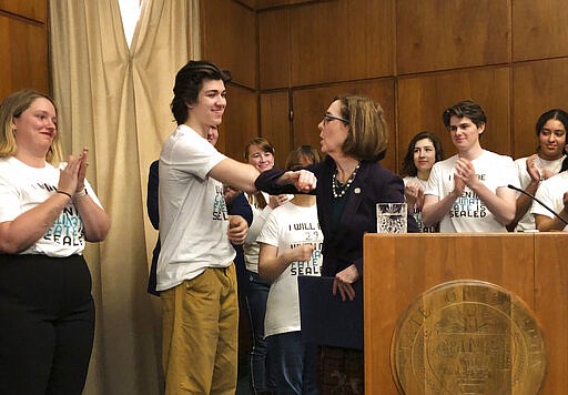 Oregon Gov. Kate brown elbow-bumps student Charlie Abrams at a ceremony on Tuesday, March 10, 2020, where she signed a sweeping executive order for the state to reduce carbon emissions to combat global warming. Amid a spreading coronavirus outbreak, many people are no longer shaking hands and are coming up with alternatives, like the elbow bump. (AP Photo/Andrew Selsky)