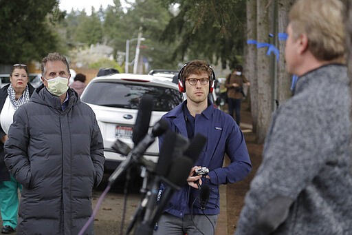 In this March 11, 2020, photo, Scott Sedlacek, left, who has tested positive for the coronavirus, wears a mask as he stands near reporters and listens to Tim Killian, right, a spokesman for Life Care Center in Kirkland, Wash., during Killian's daily briefing. Sedlacek's father Chuck, who also has tested positive, lives in the facility, which has been at the center of the coronavirus outbreak in the state. Residents of assisted living facilities and their loved ones are facing a grim situation as the coronavirus spreads across the country, placing elderly people especially at risk. (AP Photo/Ted S. Warren)