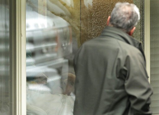 In this March 12, 2020, photo, Steve Sedlacek, right looks at his father, Chuck Sedlacek, back left, through reflections in the window of his room at Life Care Center in Kirkland, Wash., near Seattle. The facility has been at the center of the coronavirus outbreak in the state, and Sedlacek's brother Scott said that he and his siblings have barely spoken to their father inside the center, who in addition to testing positive for the coronavirus, has blindness, neuropathy, and has difficulty using a phone, saying he is more of an &quot;inmate&quot; than a patient. Residents of assisted living facilities and their loved ones are facing a grim situation as the coronavirus spreads across the country, placing elderly people especially at risk. (AP Photo/Ted S. Warren)