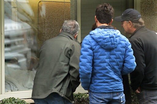 In this March 12, 2020, photo, family members of Chuck Sedlacek, left, look at him through reflections in the window of his room at Life Care Center in Kirkland, Wash., near Seattle. The facility has been at the center of the COVID-19 coronavirus outbreak in the state, and Sedlacek's son Scott said that he and his siblings have barely spoken to their father inside the center, who in addition to testing positive for the coronavirus, has blindness, neuropathy, and has difficulty using a phone, saying he is more of an &quot;inmate&quot; than a patient. Residents of assisted living facilities and their loved ones are facing a grim situation as the coronavirus spreads across the country, placing elderly people especially at risk. (AP Photo/Ted S. Warren)