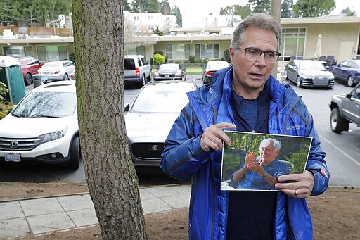 In this March 12, 2020, photo, Scott Sedlacek poses for a photo while holding a photo of his father, Chuck, outside Life Care Center in Kirkland, Wash., near Seattle. The facility has been at the center of the coronavirus outbreak in the state, and Sedlacek &#151; who also has tested positive for the virus &#151; said he and his siblings have barely spoken to their father, who in addition to testing positive for the coronavirus, has blindness, neuropathy, and has difficulty using a phone, saying he is more of an &quot;inmate&quot; than a patient. Residents of assisted living facilities and their loved ones are facing a grim situation as the coronavirus spreads across the country, placing elderly people especially at risk. (AP Photo/Ted S. Warren)