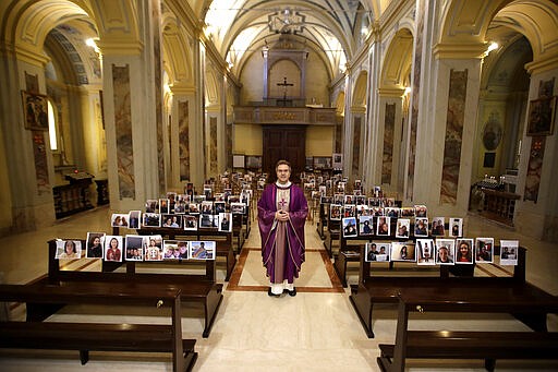 Robbiano Church parson, Don Giuseppe Corbari, poses next to selfies sent by his parishioners and pasted on the pews of the Robbiano church, as Masses for the faithful have been suspended following Italy's coronavirus emergency, in Giussano, northern Italy, Sunday, March 15, 2020.  For most people, the new coronavirus causes only mild or moderate symptoms. For some, it can cause more severe illness, especially in older adults and people with existing health problems. (AP Photo/Luca Bruno)