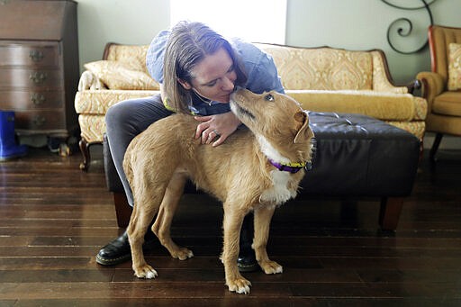 Jennifer Haller gets a kiss from her adopted foster dog, Meg, Monday, March 16, 2020, in her home in Seattle. Earlier in the day, Haller was the first person to receive a shot of a potential vaccine for COVID-19, the disease caused by the new coronavirus, at the start of the first-stage safety study clinical trial of the vaccine at the Kaiser Permanente Washington Health Research Institute in Seattle. (AP Photo/Ted S. Warren)