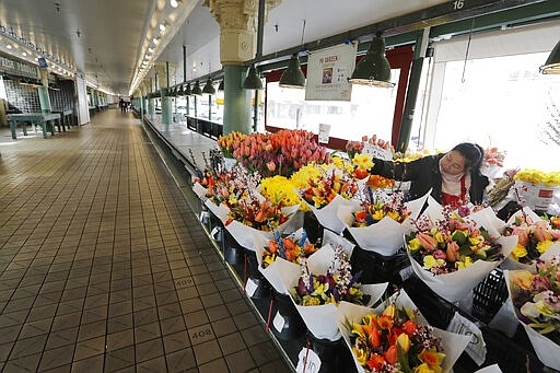 Flower vendor Shor Chang places daffodils in an arrangement while empty booths line the hall behind at the Pike Place Market Tuesday, March 17, 2020, in Seattle. Many shops in the landmark market, including fish, meat, bakeries and fruit and vegetable grocers, continue to remain open, though business has dropped considerably in recent weeks. A day earlier, Washington Gov. Jay Inslee ordered all bars, restaurants, entertainment and recreation facilities to temporarily close to fight the spread of COVID-19. (AP Photo/Elaine Thompson)