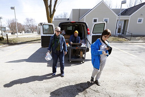 In this Tuesday, March 17, 2020, photo Des Moines Area Religious Council food pantry volunteer Louise Minor, left, carries a bag of food for Marlis Evans, of Des Moines, Iowa, right, at a senior center in Des Moines, Iowa. Amid concerns the new coronavirus has left many people to at least temporarily lose their jobs, food banks and pantries across the U.S. are scrambling to meet an expected surge in demand even as older volunteers have been told to stay home and calls for social distancing are complicating efforts to package and distribute food. (AP Photo/Charlie Neibergall)