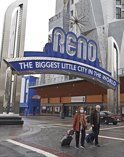 Ted and Chris Champman make their way across the street past the arch in downtown Reno, Nev., after leaving their casino hotel Wednesday, March 18, 2020. The Florida couple was planning on staying in Reno two days, but because the casino where they were staying was closing, they had to leave after their first night. Gov. Steve Sisolak issued a sweeping statewide order aimed to stop the spread of the new coronavirus.  (Andy Barron/The Reno Gazette-Journal via AP)