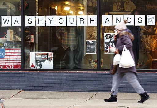 In this March 19, 2020, photo, a woman in a face mask walks past a &quot;Wash Your Hands&quot; sign after buying groceries in Oak Park, Ill., before a two-week &quot;shelter in place&quot; order goes into effect. Leaders in the Chicago suburb were the first in the state to take the step, requiring non-essential businesses to close, to try to curb the spread of the coronavirus. (AP Photo/Martha Irvine)