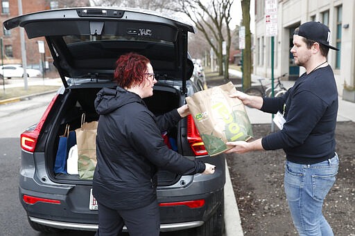 Sugar Beets Food Co-Op employee Jamie Melendy, right, gives curb service to Lola Wright in the Village of Oak Park, Ill., Friday, March 20, 2020. There are at least three confirmed cases of COVID-19 in Oak Park, just nine miles from downtown Chicago, where the mayor has ordered residents to shelter in place. With so few tests available, surely there are others, says Tom Powers, spokesman for the village of about 52,000 in a metropolitan area with millions. (AP Photo/Charles Rex Arbogast)