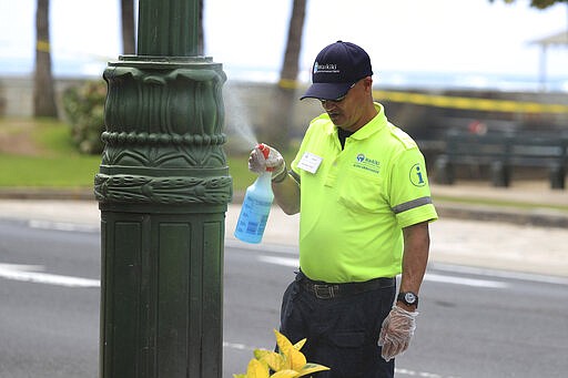 A worker sprays sanitizer on a light post Friday, March 20, 2020, in the Waikiki area of Honolulu. Honolulu closed all public parks and recreation areas Friday until the end of April in an effort to help stop the spread of the coronavirus. (AP Photo/Marco Garcia)