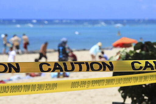 As beachgoers are seen in the background, yellow caution tape wrapped across Waikiki, Friday, March 20, 2020, in Honolulu. Honolulu closed all public parks and recreation areas Friday until the end of April in an effort to help stop the spread of the coronavirus. Many beachgoers simply went under the security tape to access the beach and shoreline. (AP Photo/Marco Garcia)