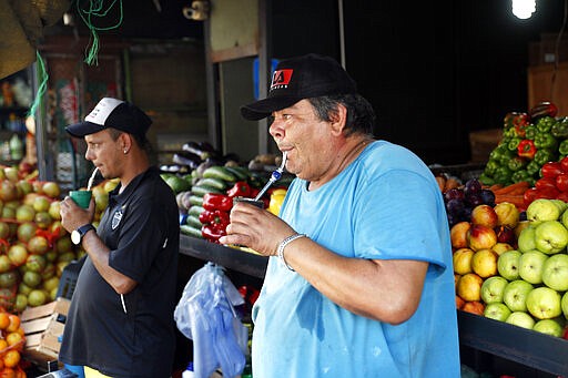 In this March 17, 2020 photo, Artemio Lezcano, front, and a work colleague, drink their own mugs of cold &quot;mate,&quot; instead of sharing one as they usually do, at their fruit stand at Mercado 4 in Asuncion, Paraguay. &quot;This is a solution to continue drinking&quot; said Lezcano.&quot;  In Uruguay, Paraguay and Argentina, drinking and sharing the herb &quot;mate&quot; is a tradition among family members, office colleagues and schoolmates, who share the straw as they pass the drink around. Today, however, the new coronavirus outbreak has made people quit sharing, to prevent spreading COVID-19.  (AP Photo/Jorge Saenz)