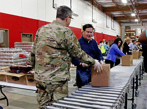 Arizona National Guard commander Major Gen. Michael McGuire, Gov. Doug Ducey, center, and Secretary of State Kimberly Yee pack food boxes at St. Mary's Food Bank in Phoenix Friday, March 20, 2020. The governor called out the Guard to help grocery stores restock shelves and went to the food bank to highlight shortages of donations and volunteers for charity operations triggered by the coronavirus outbreak. (AP Photo/Bob Christie)