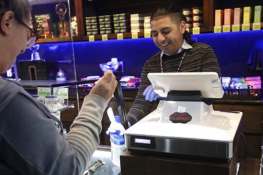 Max de Leon, right, a budtender at The Green Cross cannabis dispensary, smiles while handing a cleaning wipe to a customer in San Francisco, Wednesday, March 18, 2020. As about 7 million people in the San Francisco Bay Area are under shelter-in-place orders, only allowed to leave their homes for crucial needs in an attempt to slow virus spread, marijuana stores remain open and are being considered &quot;essential services.&quot; (AP Photo/Jeff Chiu)