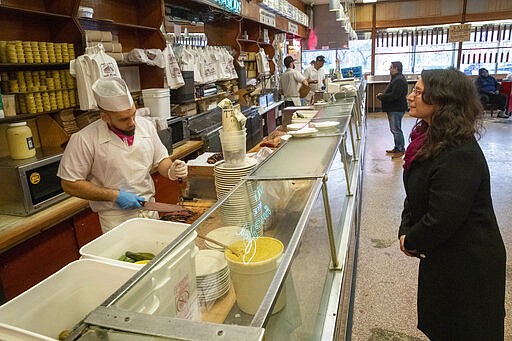 In this March 19, 2020 photo,  Adar Beck, right, of Jersey City, N.J., stands at a distance from the counter as she watches a cutter carve her order of pastrami at Katz's Delicatessen on the Lower East Side of New York. The iconic eatery is only open for take out and delivery orders due to the coronavirus outbreak. (AP Photo/Mary Altaffer)