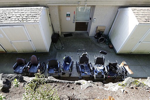 A line of wheelchairs sit outside a back door at the Life Care Center, where at least 30 coronavirus deaths have been linked to the facility, Wednesday, March 18, 2020, in Kirkland, Wash. Staff members who worked while sick at multiple long-term care facilities contributed to the spread of COVID-19 among vulnerable elderly in the Seattle area, federal health officials said Wednesday. (AP Photo/Elaine Thompson)