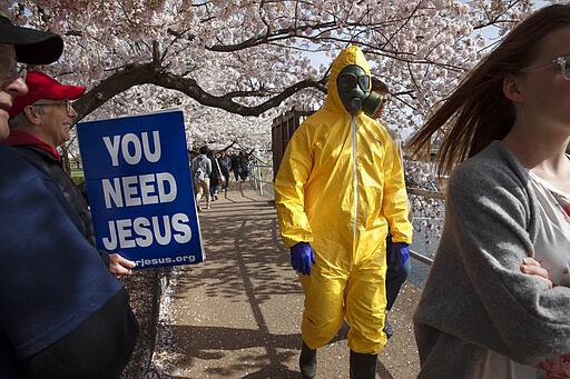 A 17-year-old who asked not to be named, wears a hazmat suit, gas mask, boots, and gloves as he walks past people holding a sign that says, &quot;you need Jesus&quot; as he and his family from Gaithersburg, Md. walk under cherry blossom trees in full bloom along the tidal basin, Sunday, March 22, 2020, in Washington. &quot;I'm not worried for me since I'm young,&quot; says the 17-year-old, &quot;I'm wearing this in case I come into contact with anyone who is older so that I won't be a threat to them.&quot; He plans to wear his protective outfit for coronavirus each time he leaves the house. Sections of the National Mall and tidal basin areas have been closed to vehicular traffic to encourage people to practice social distancing and not visit Washington's iconic cherry blossoms this year due to coronavirus concerns. The trees are in full bloom this week and would traditionally draw a large crowd. (AP Photo/Jacquelyn Martin)