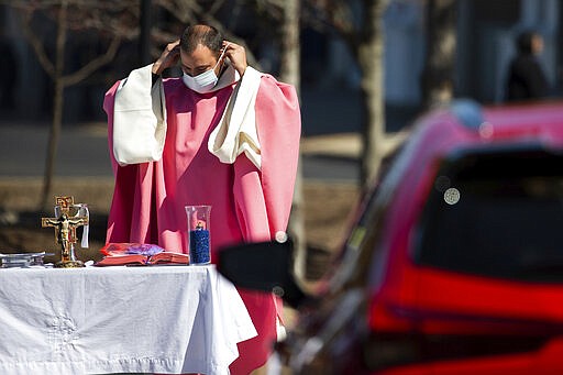 Rev. William A. Mentz, pastor of the Scranton, Pa. based St. Francis and Clare Progressive Catholic Church, puts on a mask before distributing prepackaged communion to the faithful attending mass while sitting in their cars in a parking lot at the Shoppes at Montage in Moosic, Pa. on Sunday, March 22, 2020. The Progressive Catholic Church is a small denomination operating independently of the Roman Catholic Church. Other Catholic churches in the Scranton area suspended the celebration of mass to help control the spread of COVID-19. (Christopher Dolan/The Times-Tribune via AP)