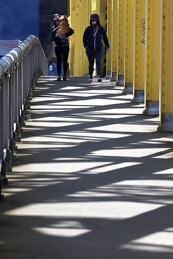 A woman carries her dog as she walks across the 16th bridge with a friend, on a sunny afternoon in downtown Pittsburgh, Sunday, March 22, 2020. (AP Photo/Gene J. Puskar)