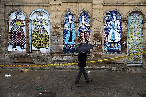 A man shelters from the rain with an umbrella a he walks past an old building decorated with replica of Iranian old paintings in a mostly empty street in a commercial district in downtown Tehran, Iran, Sunday, March 22, 2020. On Sunday, Iran imposed a two-week closure on major shopping malls and centers across the country to prevent spreading the new coronavirus. Pharmacies, supermarkets, groceries and bakeries will remain open. (AP Photo/Vahid Salemi)