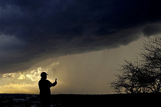 In this March 19, 2020, photo, a man looks at his phone storm clouds pass overhead from the Liberty Memorial in Kansas City, Mo. The coronavirus pandemic is leading to information overload for many people, often making it difficult to separate fact from fiction and rumor from deliberate efforts to mislead. (AP Photo/Charlie Riedel)