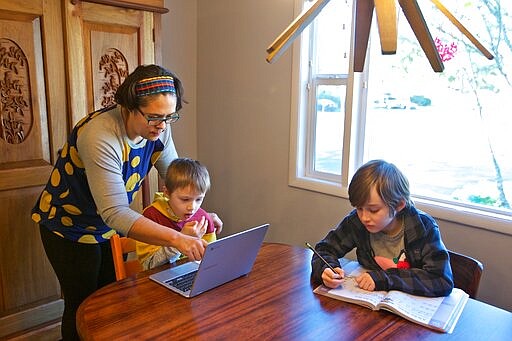 In this March 17, 2020 photo Olivia Bucks, left, helps her son Keith Bucks, center, with an online class assignment while Ashton Morris, right, works on a handwriting lesson from their first grade class at Arco Iris Spanish Immersion School in Beaverton, Ore. Bucks works from home selling books online and now spends her time between working on her business and helping her sons with their school work. They are using her work laptop to access their classroom assignments. (AP Photo/Craig Mitchelldyer)