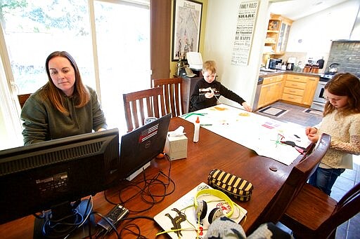 In this Tuesday, March 17, 2020 photo Kim Borton, left, works from home while her children Logan Borton, center, age 6 and Katie Borton, age 7, as they work on an art project in Beaverton, Ore. Borton works for Columbia Sportswear in supply chain account operations. Her children attend Hiteon Elementary school and have sent home some home work packets and emails with links for remote learning, but she has also added her own curriculum to their day to fill the voids so she can continue to work and keep the kids busy. (AP Photo/Craig Mitchelldyer)